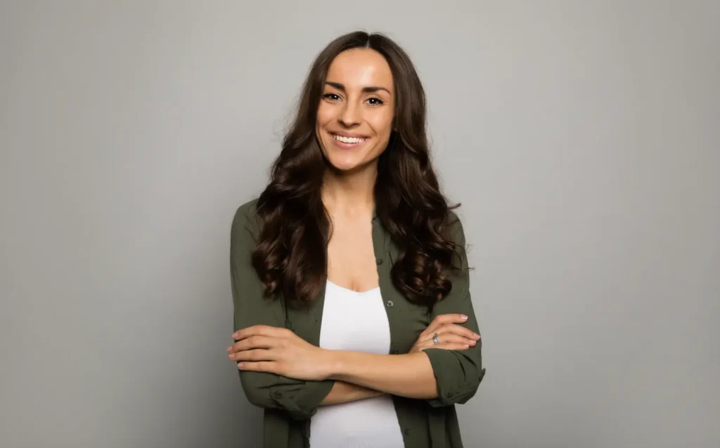 Smiling woman with long dark hair stands with her arms crossed against a gray background.